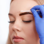 Cosmetologist cleaning face of client during permanent make up of eyebrows, using cotton buds . Professional making procedure in blue gloves for client in beauty salon.